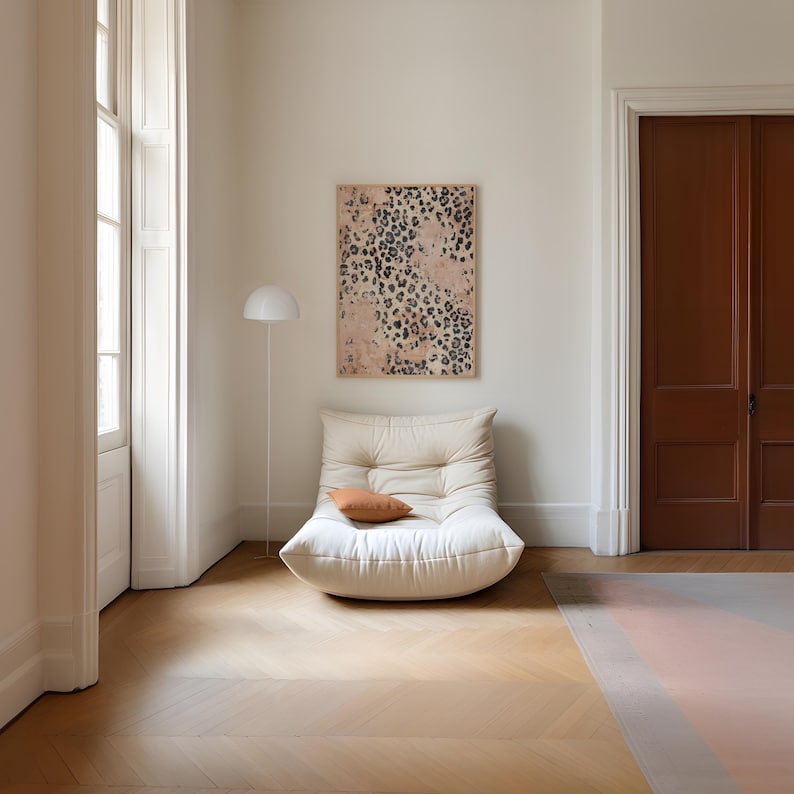 May include: A white, tufted, oversized armchair with a brown throw pillow sits in a room with white walls and hardwood floors. A white lamp stands in the corner of the room. A framed print with a pink and black animal print design hangs on the wall.