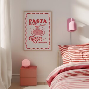 May include: A white poster with red text that reads "PASTA IS MY LOVE LANGUAGE" with a stylized illustration of a plate of spaghetti and a fork. The poster is hanging on a white wall above a red nightstand with a pink lamp.