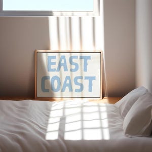 May include: A framed print with the words "EAST COAST" in light blue lettering on a white background. The print is sitting on a white bed with a white pillow.