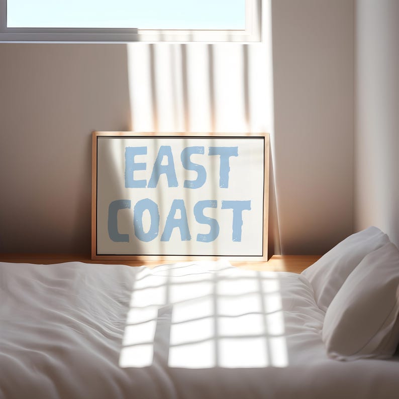 May include: A framed print with the words "EAST COAST" in light blue lettering on a white background. The print is sitting on a white bed with a white pillow.