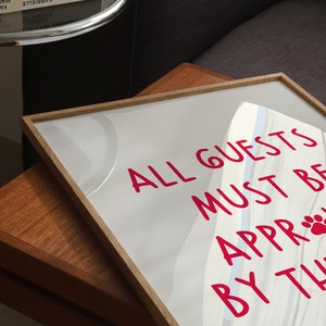 Puede incluir: Un cartel blanco enmarcado con un mensaje rosa que dice "ALL GUESTS MUST BE APPROACHED BY THE PAW". El cartel est&aacute; sobre una mesa de madera.