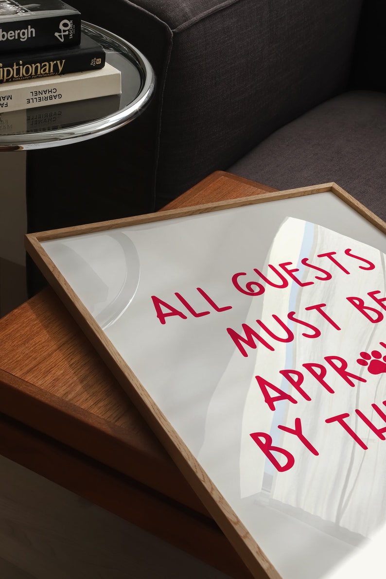 Puede incluir: Un cartel blanco enmarcado con un mensaje rosa que dice "ALL GUESTS MUST BE APPROACHED BY THE PAW". El cartel est&aacute; sobre una mesa de madera.