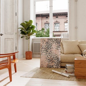 May include: A living room interior with a large canvas print featuring a pink and black leopard print design. The print is hanging on a white wall above a beige sofa. A potted ficus tree is in the corner of the room.