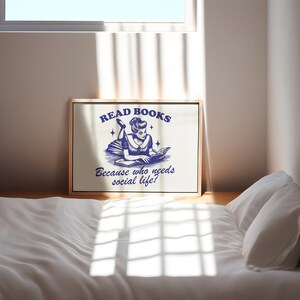 May include: A framed print with the text "READ BOOKS Because who needs social life?" in navy blue. The illustration depicts a woman reading a book. The print is displayed on a wooden floor.