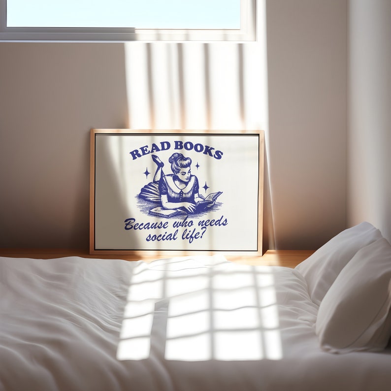May include: A framed print with the text "READ BOOKS Because who needs social life?" in navy blue. The illustration depicts a woman reading a book. The print is displayed on a wooden floor.