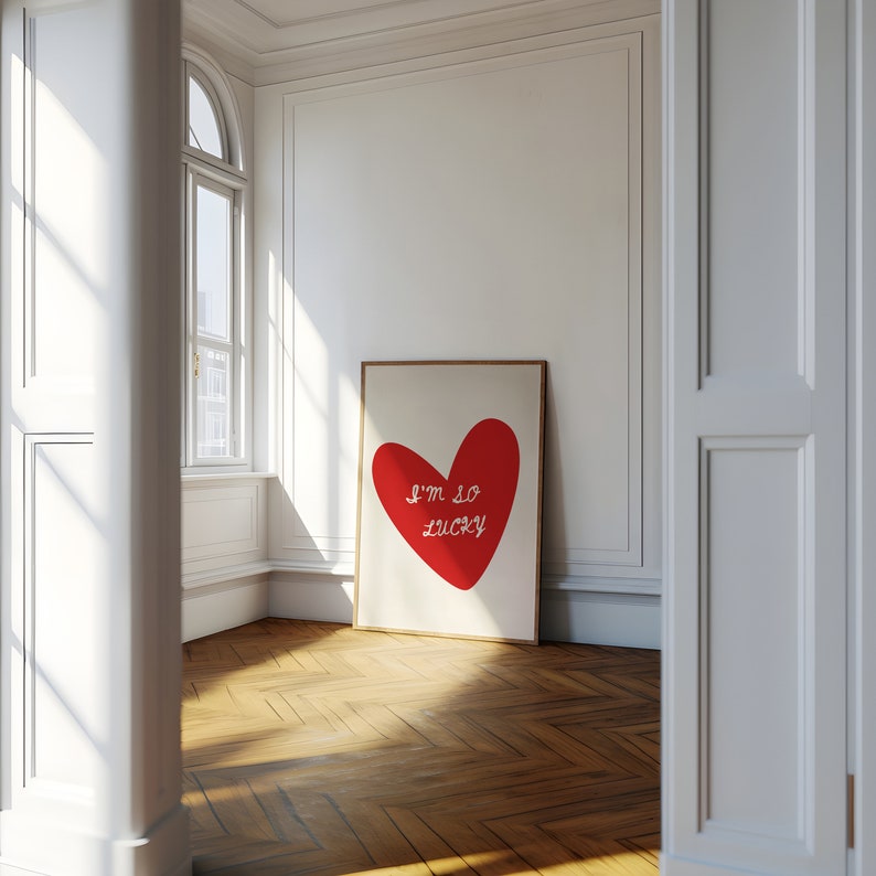 May include: A framed print with a red heart and the text "I'm so lucky" in white. The print is hanging on a white wall in a room with hardwood floors.