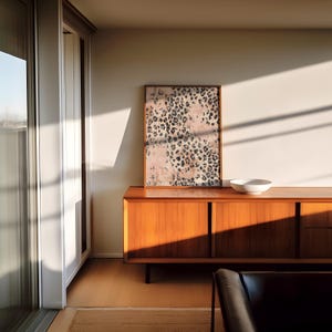 May include: A mid-century modern style wooden cabinet with three drawers and two doors. The cabinet is a light brown color and has a white ceramic bowl on top. A framed print with a pink and black animal print design hangs on the wall behind the cabinet.