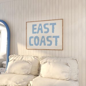 May include: A white couch with two white pillows and a framed print on the wall that says "EAST COAST" in blue lettering.