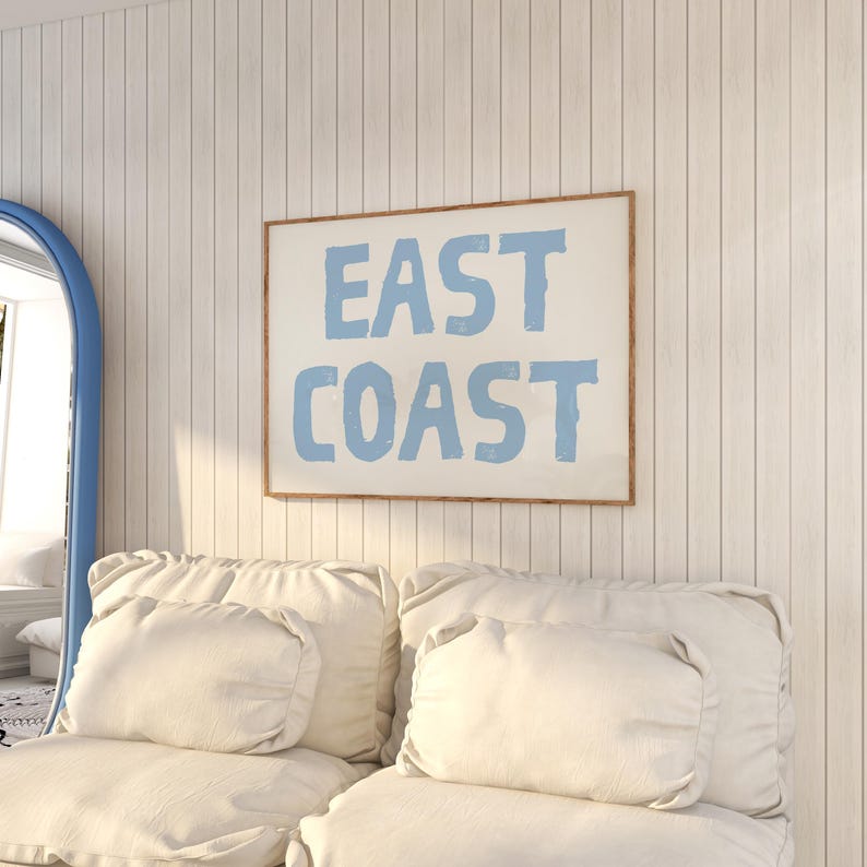 May include: A white couch with two white pillows and a framed print on the wall that says "EAST COAST" in blue lettering.