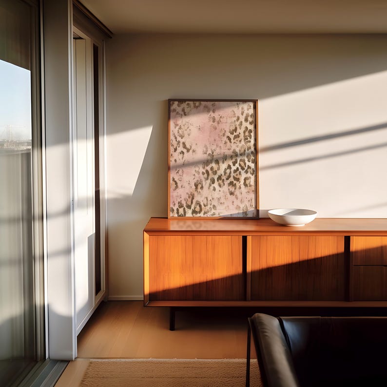 May include: A mid-century modern style wooden cabinet with three drawers and two doors. The cabinet is in a living room with a large framed print of an abstract animal print design in pink and brown tones.