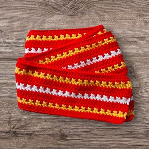 May include: A folded, handmade crochet scarf in a vibrant red color, featuring horizontal stripes of white and yellow. The scarf is made with a textured stitch, creating a visually appealing pattern. The scarf is a cozy accessory.