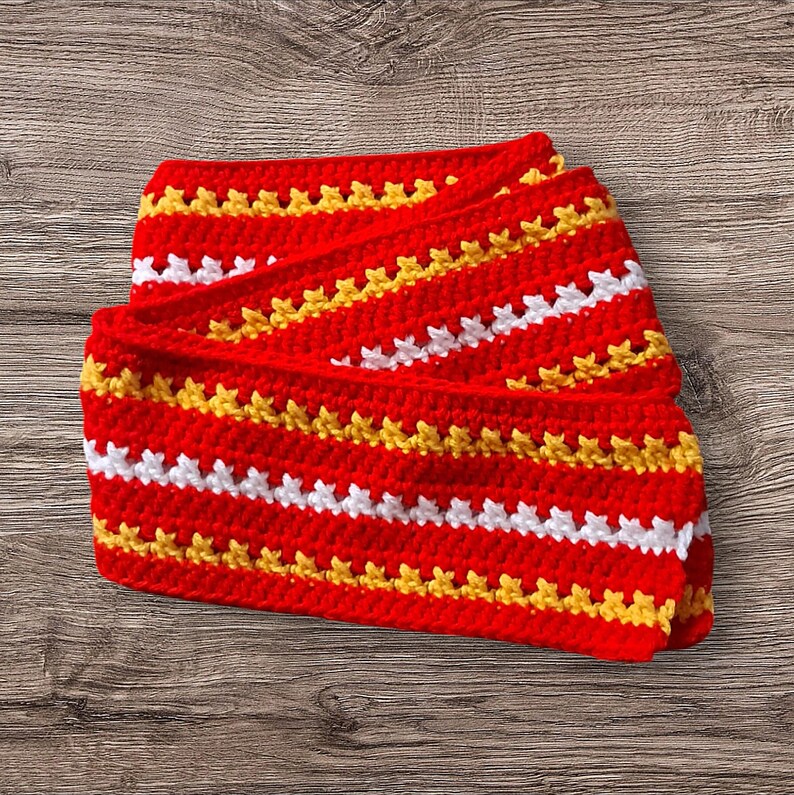 May include: A folded, handmade crochet scarf in a vibrant red color, featuring horizontal stripes of white and yellow. The scarf is made with a textured stitch, creating a visually appealing pattern. The scarf is a cozy accessory.