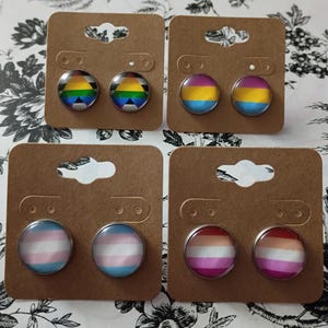 May include: Four pairs of round stud earrings on brown earring cards. Each pair features a different LGBTQ+ pride flag design: rainbow, pansexual, transgender, and lesbian. The earrings are displayed on a floral patterned background.