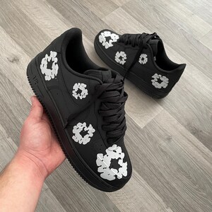 May include: Black leather low-top sneakers with white floral designs. The sneakers have a black swoosh logo on the side and the word "AIR" on the heel.