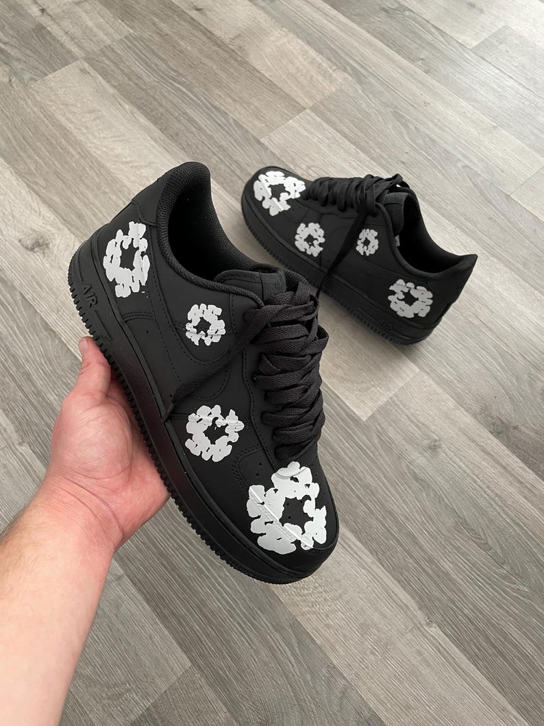 May include: Black leather low-top sneakers with white floral designs. The sneakers have a black swoosh logo on the side and the word "AIR" on the heel.