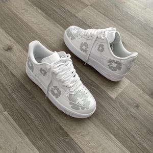 May include: A pair of white leather sneakers with a grey camouflage pattern. The sneakers have white laces and a white sole.