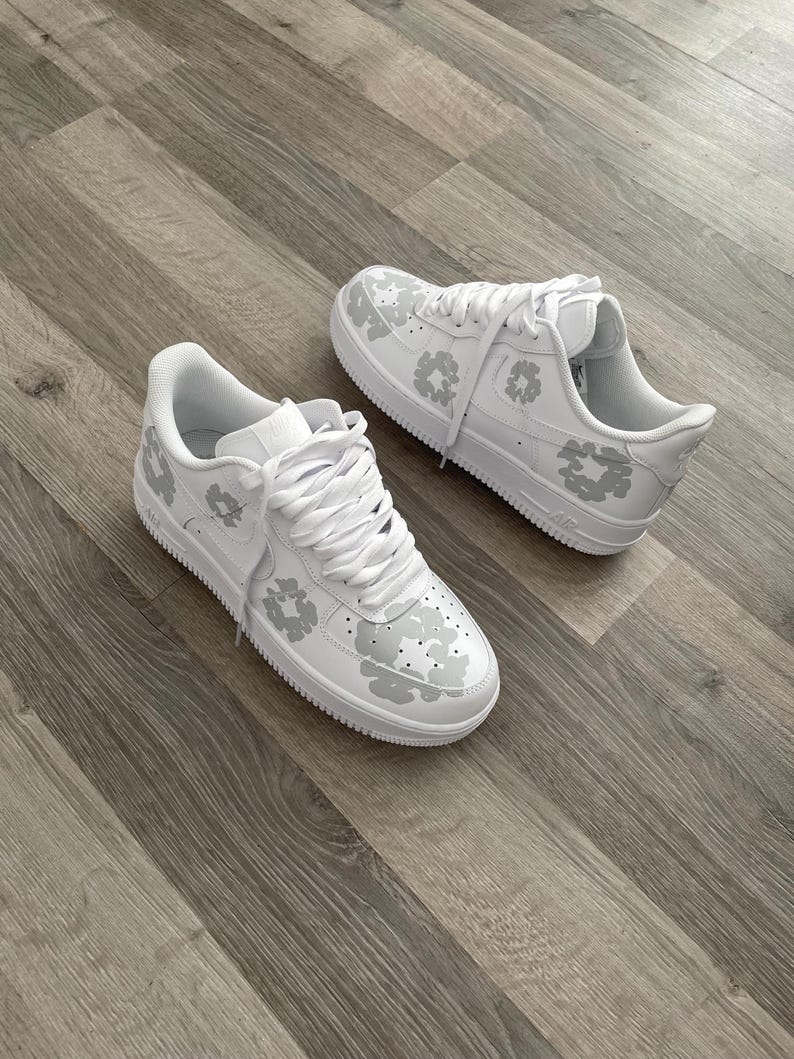 May include: A pair of white leather sneakers with a grey camouflage pattern. The sneakers have white laces and a white sole.