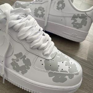 May include: A pair of white leather sneakers with a grey floral pattern. The sneakers have white laces and a white sole with the word "Air" printed on the side.