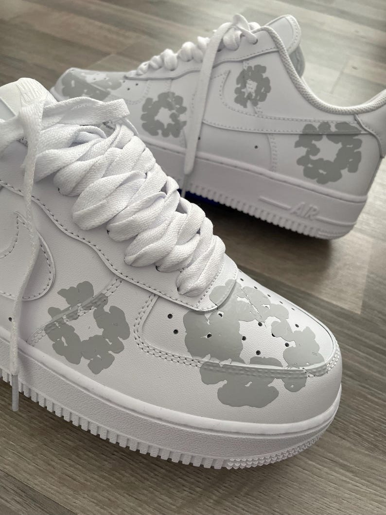 May include: A pair of white leather sneakers with a grey floral pattern. The sneakers have white laces and a white sole with the word "Air" printed on the side.