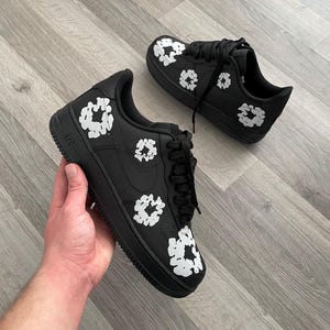 May include: Black leather sneakers with white floral designs. The sneakers have a white swoosh logo on the side and the word "AIR" on the back.
