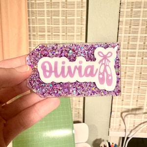 May include: A clear plastic tag with a purple glitter background and the name "Olivia" in white lettering. A purple silhouette of ballet shoes with a bow is on the right side of the tag.