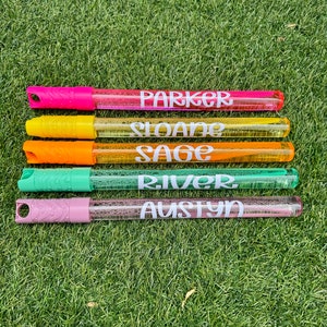 May include: Five colorful bubble wands with personalized names: Parker, Sloane, Sage, River, and Austyn. The wands are arranged vertically on a green grassy surface.