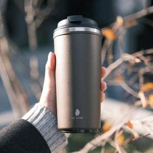 May include: A brown stainless steel travel mug with a black lid and a silver band around the top. The mug has the text "LE NOMI" printed on the side.