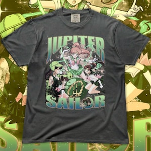 Può includere: T-shirt grigio scuro con una grafica colorata di personaggi anime e la scritta "JUPITER SAILOR" in verde e blu. La maglietta ha un aspetto vintage e invecchiato ed è realizzata in un materiale morbido.
