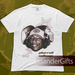 Puede incluir: Camiseta blanca con un gráfico en tonos sepia de Pharrell Williams con auriculares y gorra. El diseño incluye formas de estrella y el texto "Pharrell Williams". También es visible el nombre de la marca "ExsanderGifts".