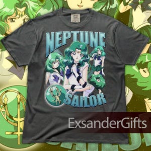May include: Dark gray t-shirt featuring a graphic of Sailor Neptune with green hair and a blue and white outfit. The text "NEPTUNE SAILOR" is displayed in blue and white. The shirt has a vintage style.