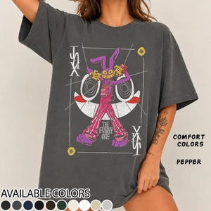 May include: A charcoal gray short-sleeve t-shirt featuring a graphic of a purple rabbit character with a wide grin, wearing pink overalls and striped pants. The design includes the text THE FUNNY ONE and abstract geometric lines in white.