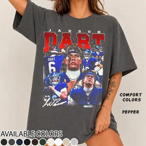 May include: A charcoal gray short-sleeve t-shirt featuring a graphic print of football player Jaxson Dart. The design includes multiple images of the player in a blue uniform, the name JAXSON DART in red block letters, and a signature. The text COMFORT COLORS and PEPPER appears on the right side.