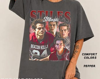 May include: A charcoal gray short-sleeve t-shirt featuring a graphic print of Stiles Stilinski from Teen Wolf. The design includes four images of the character, the text STILES STILINSKI, and BEACON HILLS 24. The shirt is labeled Comfort Colors Pepper.