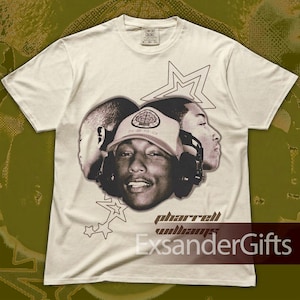 May include: Off-white t-shirt with a sepia-toned graphic of a person wearing a hat and headphones, with the text "Pharrell Williams" and "ExsanderGifts" below. The shirt has a crew neck and short sleeves.