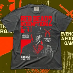 May include: Dark gray t-shirt featuring a graphic from the video game Red Dead Redemption 2. The design includes the game title in red, a portrait of Arthur Morgan, and the text "Outlaws for Life."