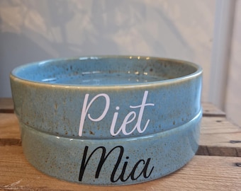 May include: Two stacked, light blue ceramic bowls with a speckled texture. The top bowl has the name "Piet" in white script, and the bottom bowl has "Mia" in black script. The bowls are sitting on a wooden surface.