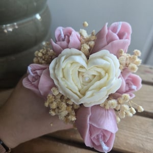 Könnte beinhalten: Ein handgemachter Seifenblumenstrauß. Die Anordnung zeigt rosa Rosenknospen, die eine herzförmige cremefarbene Rose umgeben. Kleine, getrocknete, hellbraune Füllblumen sind im ganzen Strauß verstreut.
