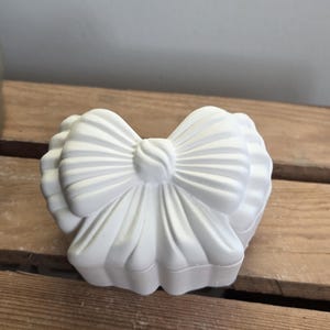 Könnte beinhalten: Eine weiße, schleifenförmige dekorative Box. Die Box hat ein detailliertes Schleifendesign auf der Oberseite, mit gerippten Details auf der Schleife und den Kanten der Box. Die Box besteht aus einem festen, undurchsichtigen Material und steht auf einer Holzoberfläche.
