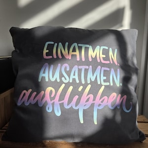 Könnte beinhalten: Ein dunkelgraues quadratisches Kissen mit den Worten "EINATMEN. AUSATMEN. ausflippen" in einem Regenbogen-Farbverlauf. Das Kissen liegt auf einer Holzoberfläche, mit Sonnenlicht und Schatten im Hintergrund.