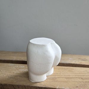 Könnte beinhalten: Ein weißer, geformter Keramik-Blumentopf in Form einer menschlichen Gestalt. Der Topf hat eine abgerundete Form mit zwei kleinen Füßen. Die Oberfläche hat ein strukturiertes Aussehen. Der Topf steht auf einer Holzoberfläche.