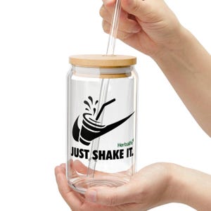 Puede incluir: Un tarro de vidrio transparente para beber con tapa de madera y pajita transparente. El tarro tiene un dise&ntilde;o gr&aacute;fico negro con las palabras "JUST SHAKE IT." y el logotipo de Herbalife. El tarro lo sostiene una persona.