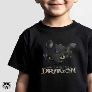 Puede incluir: Camiseta negra con un gráfico detallado de Desdentado de "Cómo entrenar a tu dragón". El dragón tiene ojos verdes y una apariencia cosida. El texto "HOW TO TRAIN YOUR DRAGON" está debajo de la imagen del dragón.