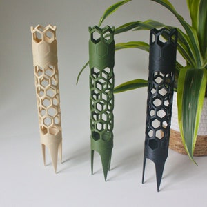 Op de afbeelding: Drie plantenondersteuningen van plastic, elk in een andere kleur: beige, groen en zwart. De ondersteuningen zijn honingraatvormig en hebben drie punten aan de onderkant.