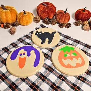 May include: Three Halloween-themed coasters with a black cat, a ghost, and a jack-o'-lantern design. The coasters are round and made of a beige material.