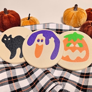 Könnte beinhalten: Drei runde Untersetzer mit Halloween-Motiven. Der erste Untersetzer zeigt eine schwarze Katze, der zweite einen lila Geist und der dritte einen orangefarbenen Kürbis mit einem grünen Stiel.