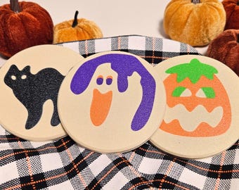 May include: Three round coasters with Halloween-themed designs. The first coaster features a black cat, the second a purple ghost, and the third an orange pumpkin with a green stem.