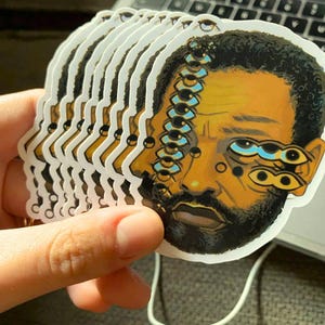 May include: A stack of die-cut vinyl stickers featuring a stylized illustration of a person with a dark beard and short hair. The artwork depicts a surreal face with multiple eyes arranged in a vertical line and clustered near the cheek area.