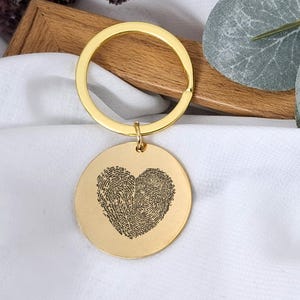 Könnte beinhalten: Ein goldfarbener Schlüsselanhänger mit einem runden Anhänger mit einem Fingerabdruck-Herz-Design. Der Schlüsselanhänger hat einen großen kreisförmigen Ring und einen kleineren Ring, der den Anhänger verbindet. Der Hintergrund umfasst einen Holzrahmen und weißen Stoff.