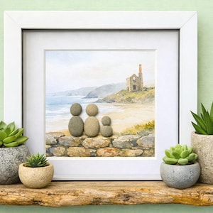 May include: A framed artwork featuring a watercolour coastal scene with a stone engine house on a cliff. Three pebble figures sit on a stone wall in the foreground. The frame is white and square, displayed with four small potted succulent plants.