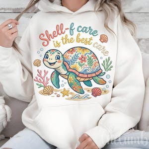 Può includere: Felpa con cappuccio color crema con un design a forma di tartaruga dei cartoni animati. La tartaruga ha un guscio a fantasia floreale ed è circondata da conchiglie, coralli e la scritta "Shell-f care is the best care". Il design utilizza colori blu, verde, giallo e rosa.